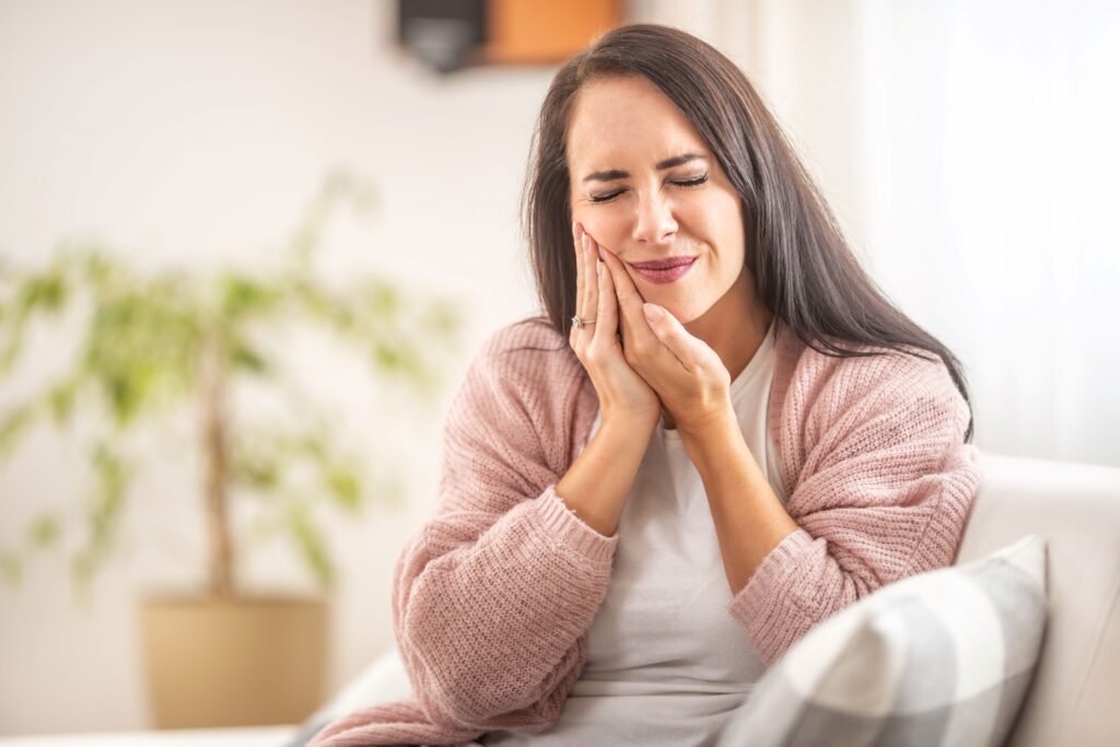 Woman in pink sweater on couch pressing 2 hands to jaw in pain